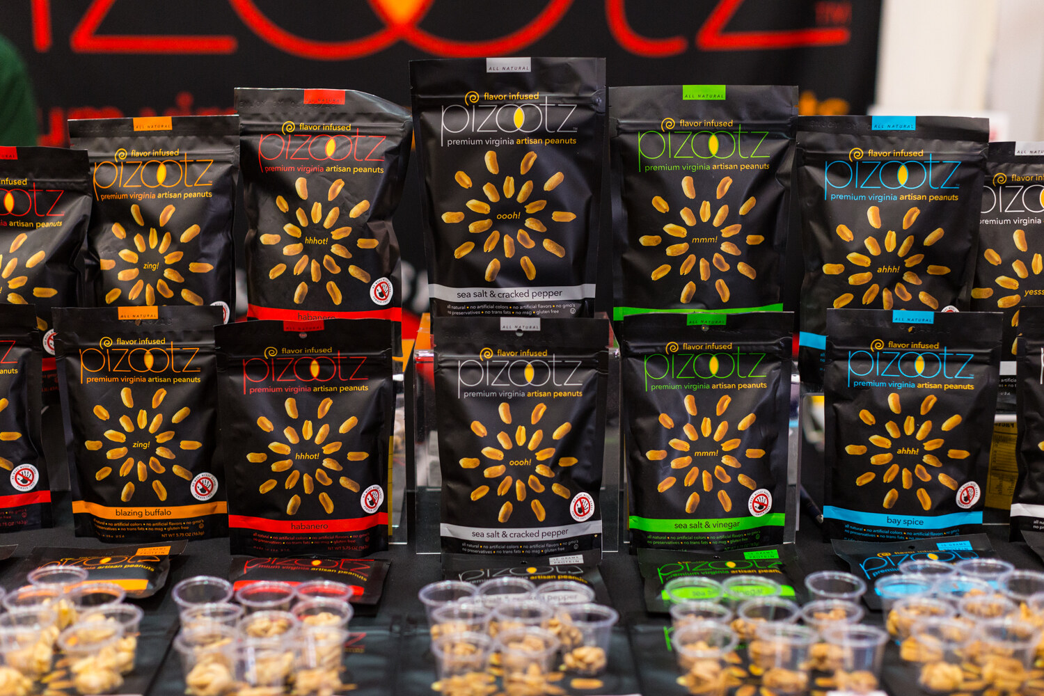 Rows of black Pizootz peanut bags with colored flavor bands above small plastic cups of peanut samples.