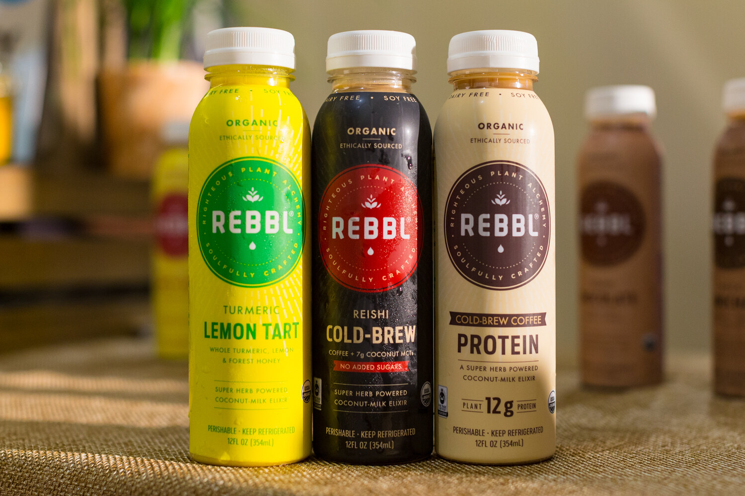 Three Rebbl bottles in yellow, black and cream on a burlap table with more bottles blurred behind.