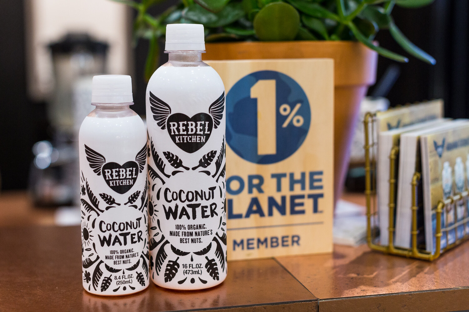 Two white bottles with black leaf and heart artwork labeled Rebel Kitchen Coconut Water next to a wooden 1% for the Planet Member sign.