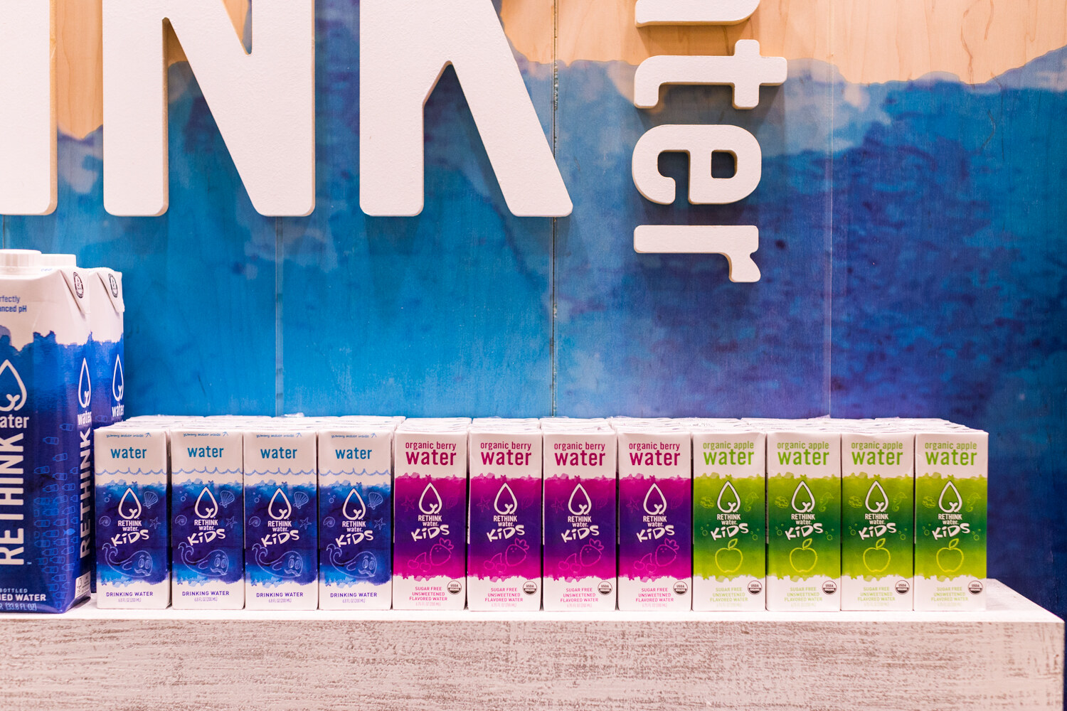 Rows of blue, purple and green Rethink Water Kids cartons on a shelf beneath a watercolor blue backdrop with large white letters.