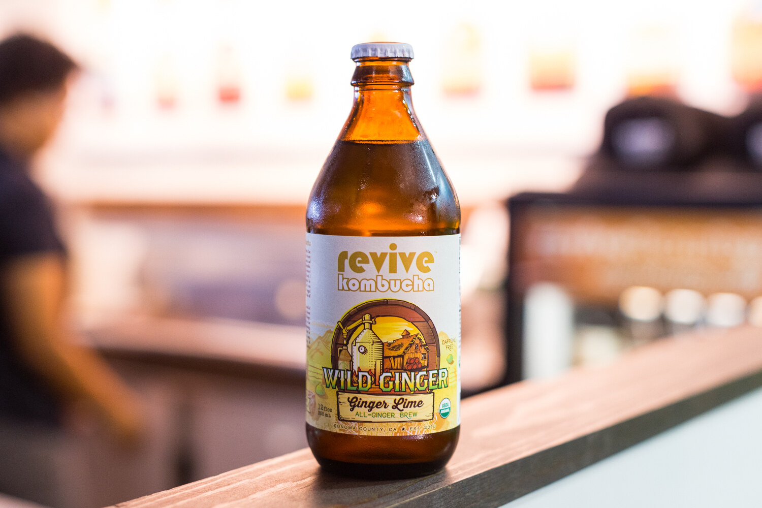A single amber bottle of Revive Kombucha Wild Ginger Ginger Lime on a wooden ledge with a blurred booth background.