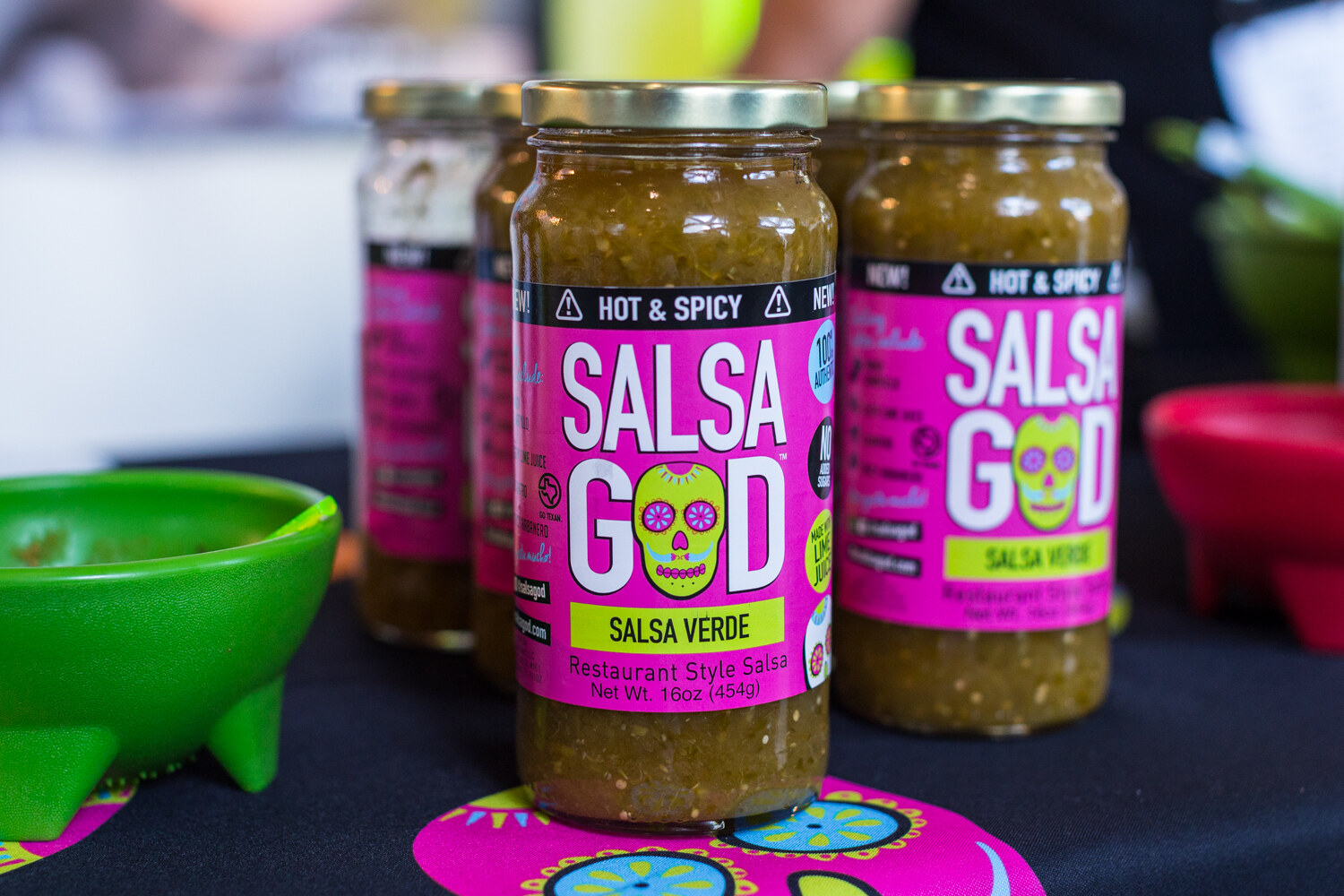 Pink-labeled Salsa God Salsa Verde jars with a skull logo on a black tablecloth printed with colorful skulls.