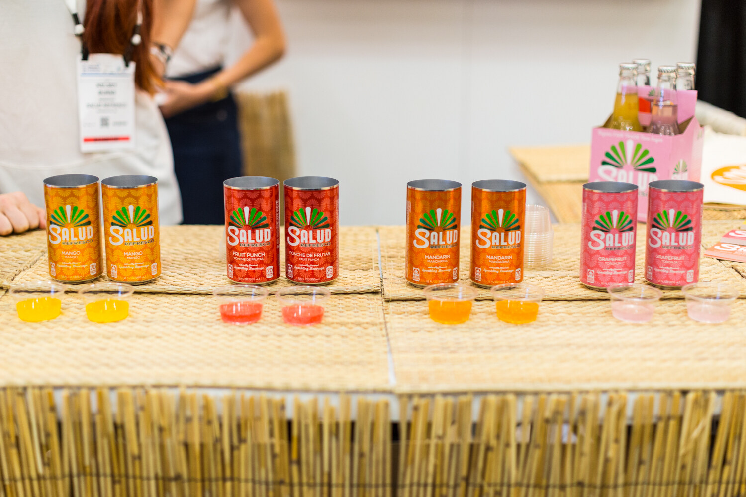 Pairs of orange, red and pink Salud cans on a woven counter with small sample cups of juice in front and an exhibitor partly visible behind.