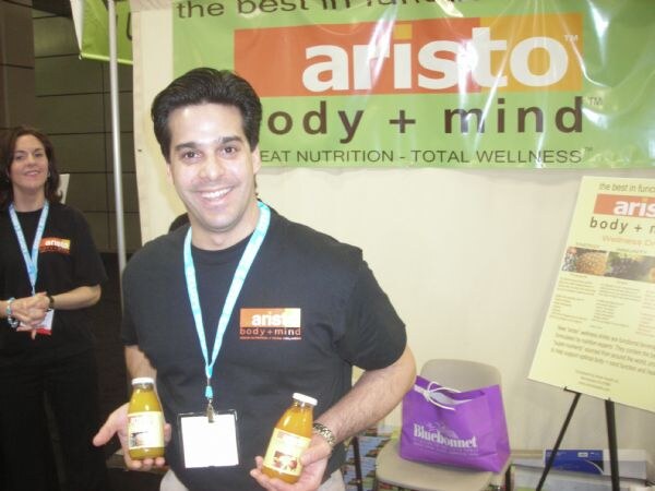 A smiling man in a black Aristo body + mind shirt holds two bottles of juice at the Aristo booth. A woman stands behind him, and a purple Bluebonnet bag sits on a chair.