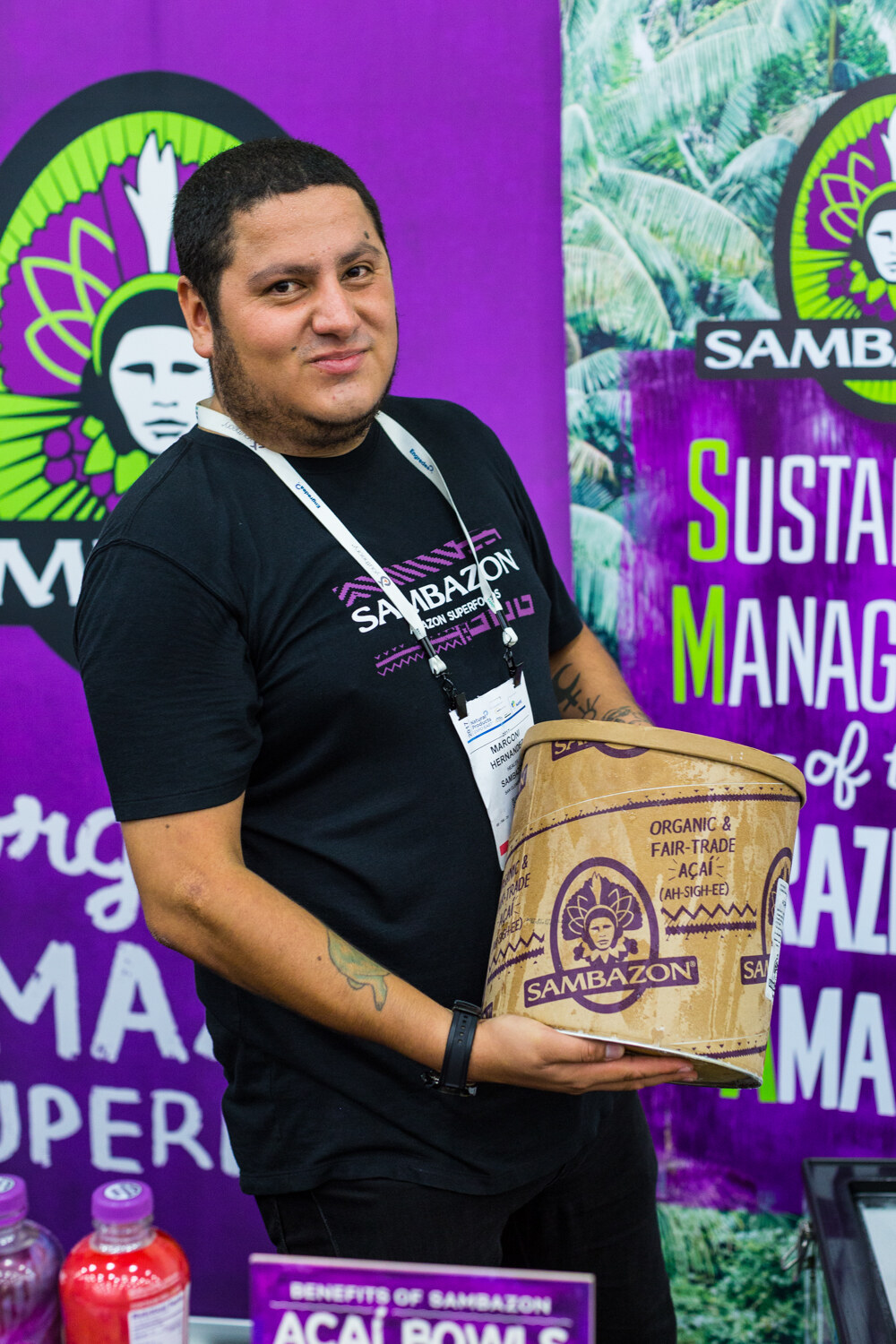 A man in a black Sambazon t-shirt and lanyard holds a paper tub of acai in front of a purple Sambazon banner.
