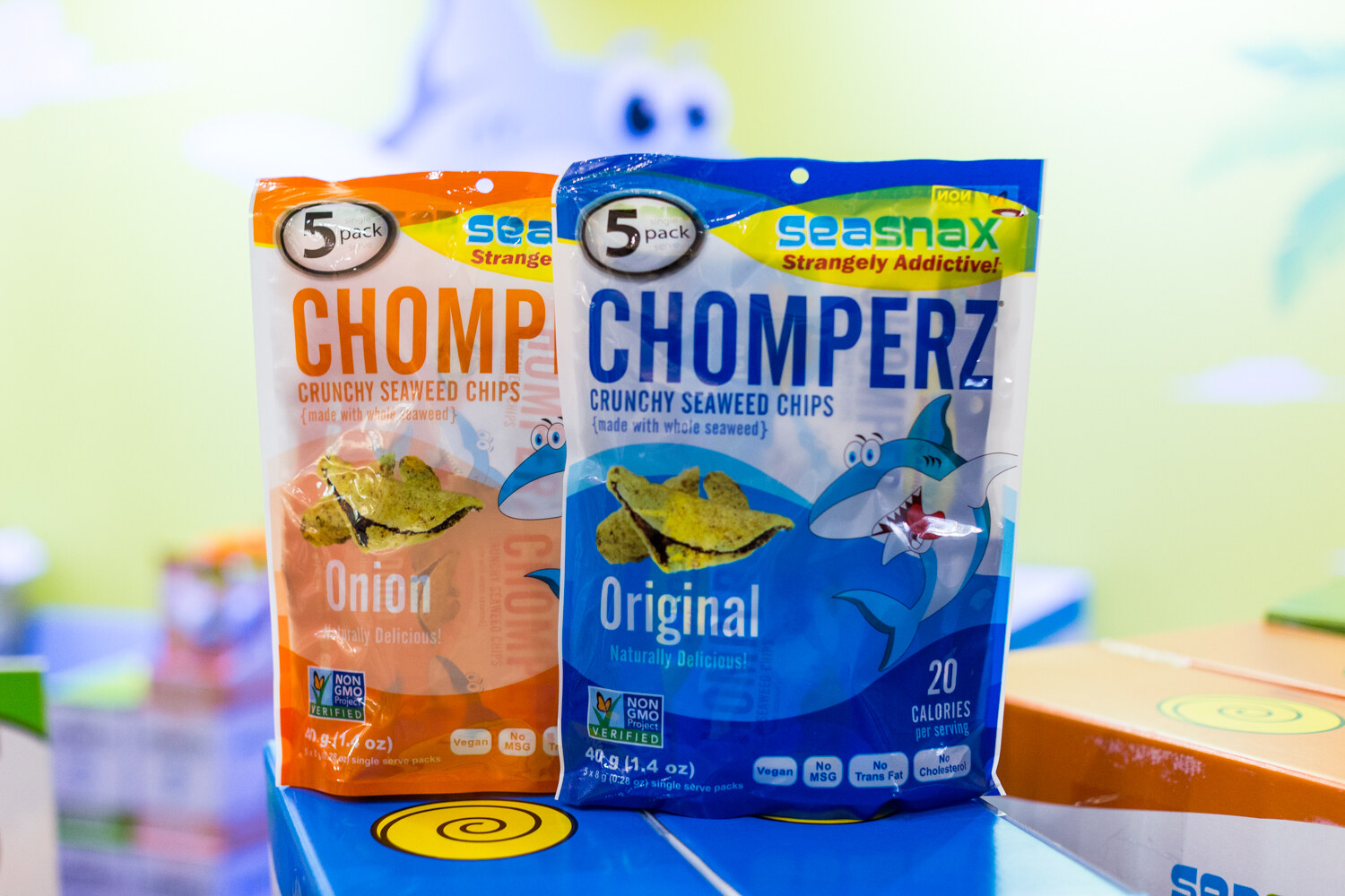 An orange and a blue bag of SeaSnax Chomperz seaweed chips with a shark cartoon, set on blue boxes.