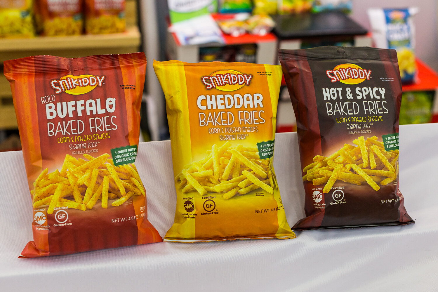 Snikiddy baked fries bags in red, yellow and dark brown standing on a white cloth with other snacks blurred behind.