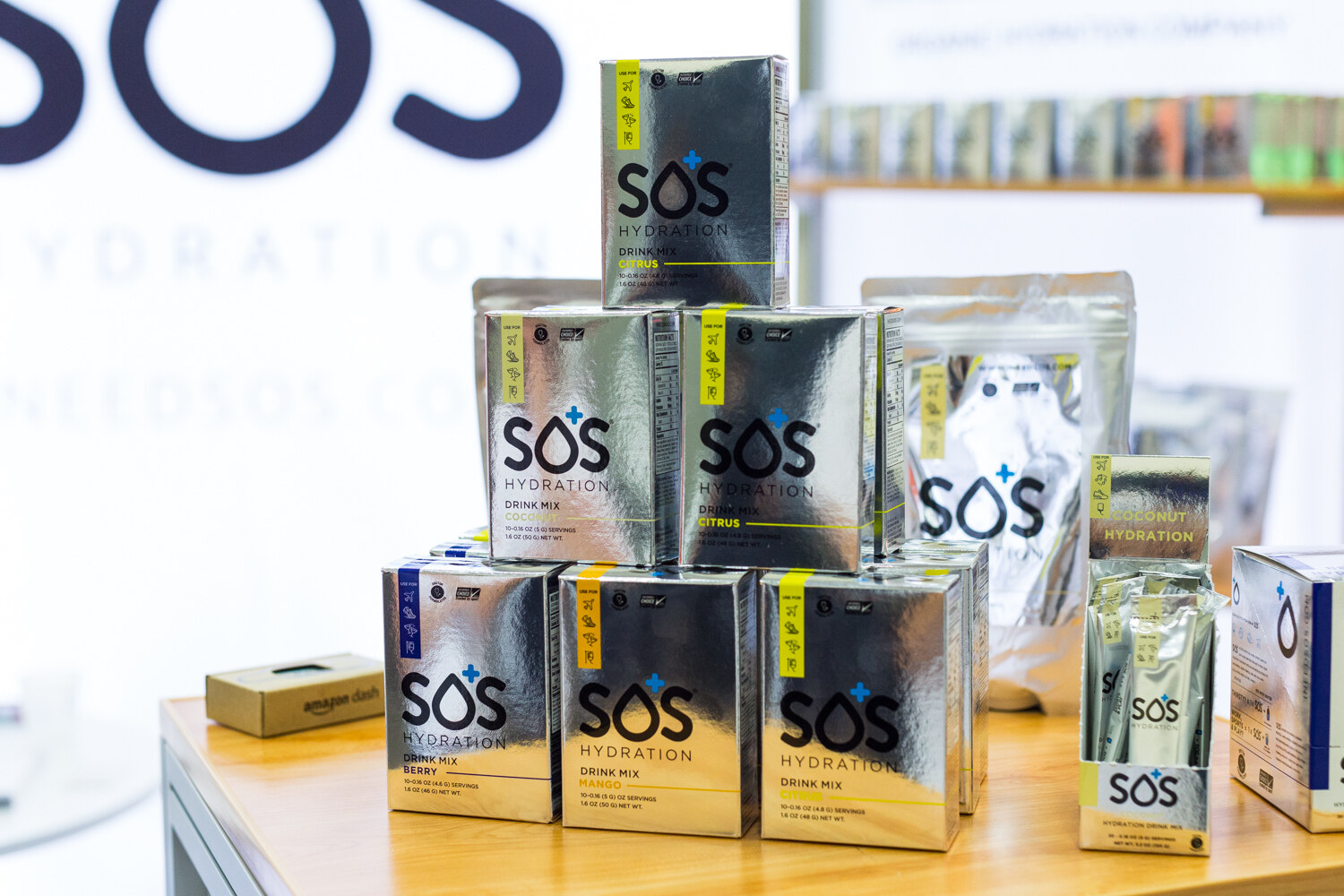A pyramid of metallic silver SOS Hydration drink mix boxes on a wooden table in front of a white SOS banner.