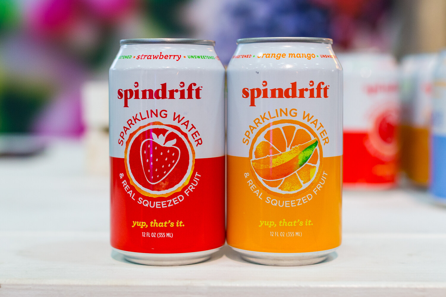 Two Spindrift sparkling water cans, one red and one orange, on a white shelf with more cans blurred behind.