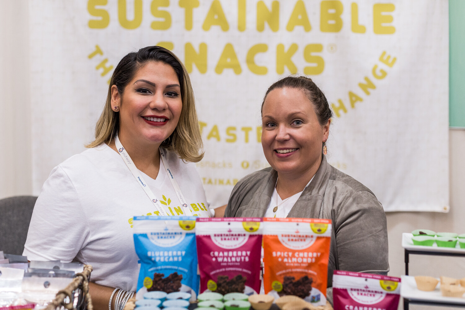 Two smiling women stand behind a table of Sustainable Snacks bags in front of a cream banner with yellow lettering.