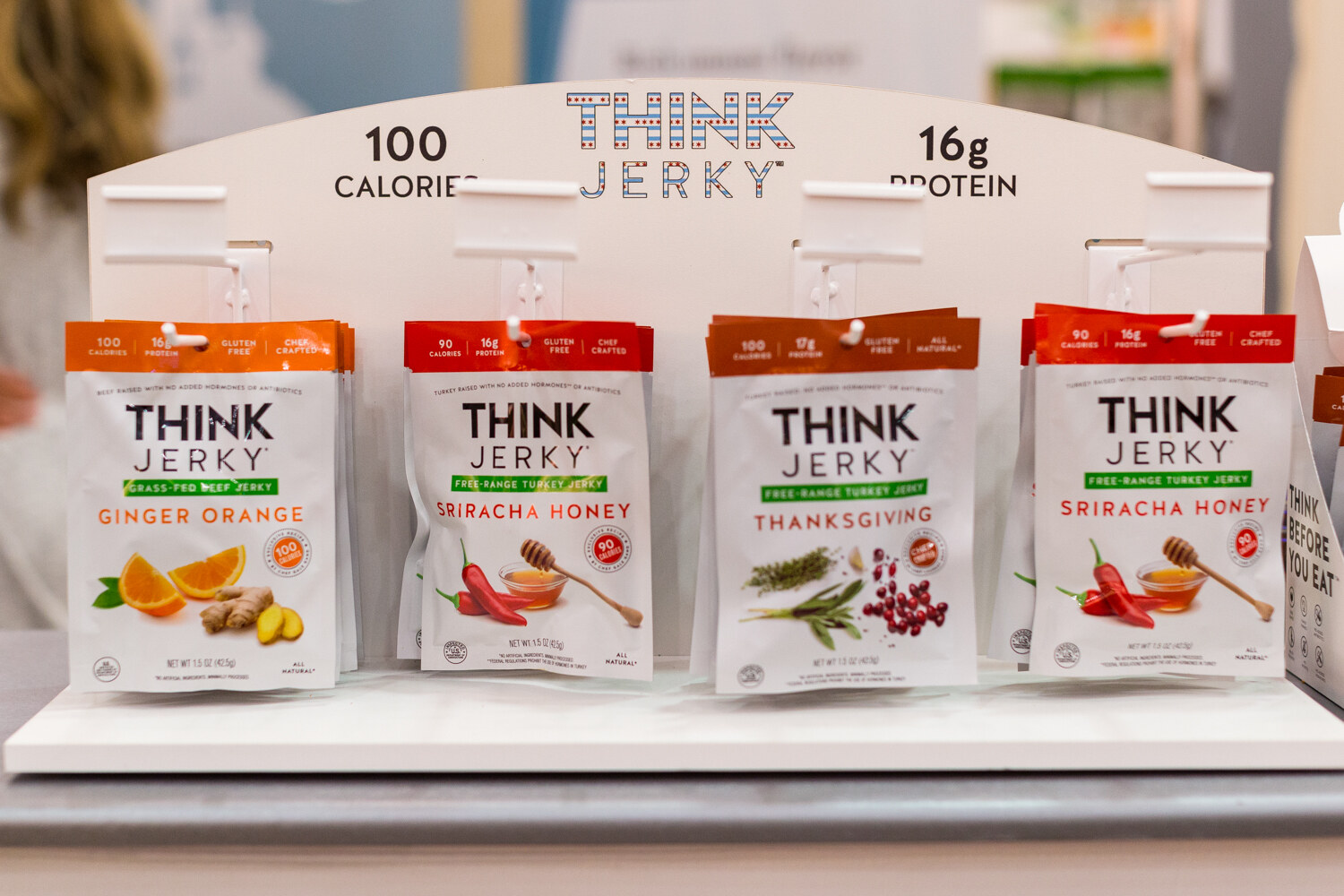Four Think Jerky pouches hanging from hooks on a white display board reading 100 Calories and 16g Protein.