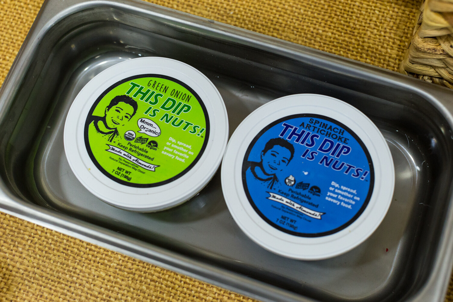 Two round tubs with green and blue lids reading This Dip Is Nuts in a steel pan on a burlap surface.