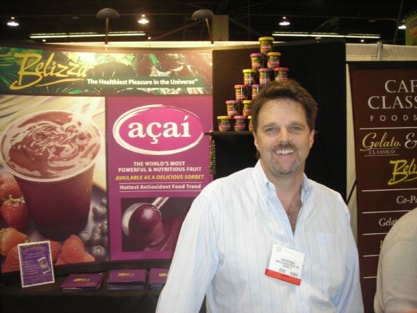 A smiling bearded man in a light striped shirt stands in front of Belizza açaí signage, with stacked sorbet cups on a shelf and a Café Classico Foods banner to the right.