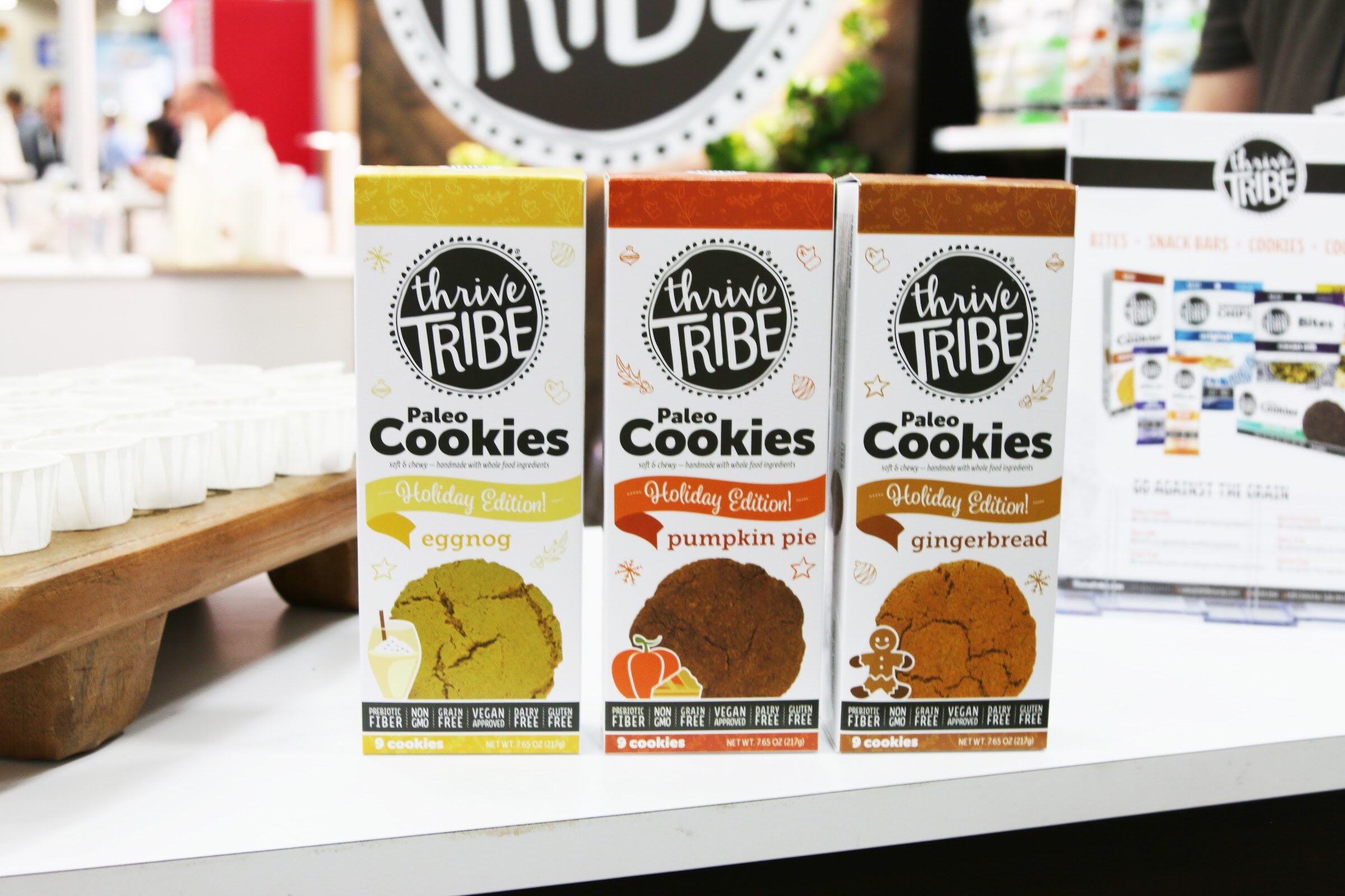 Three Thrive Tribe cookie boxes in yellow, orange and brown beside a wooden tray of paper sample cups.