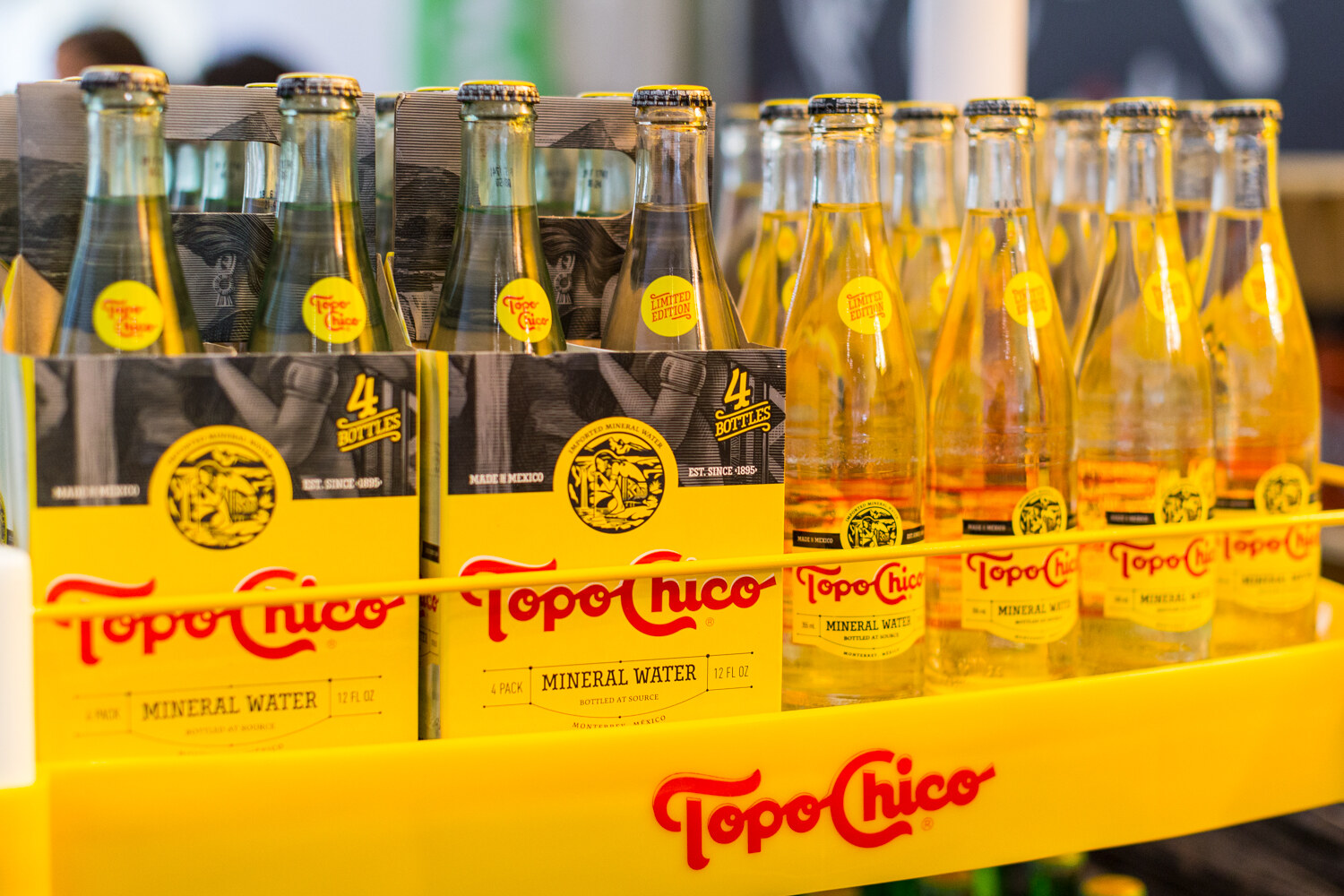Glass Topo Chico bottles with yellow Limited Edition stickers in four-pack carriers on a yellow tray with the logo.