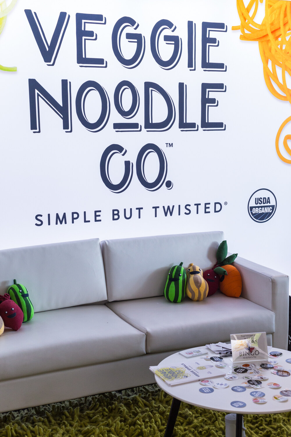 A white couch with plush vegetable toys and a small table of stickers beneath a wall reading Veggie Noodle Co. Simple But Twisted.