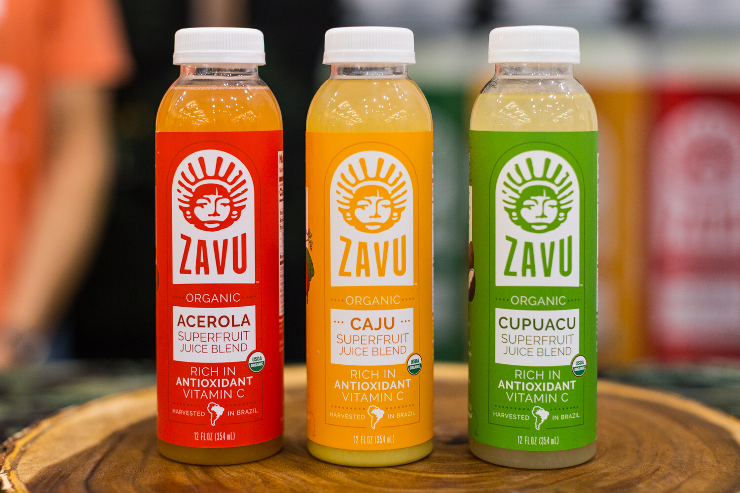 Three bottles with red, orange and green Zavu labels on a tree-slice board with a blurred booth behind.
