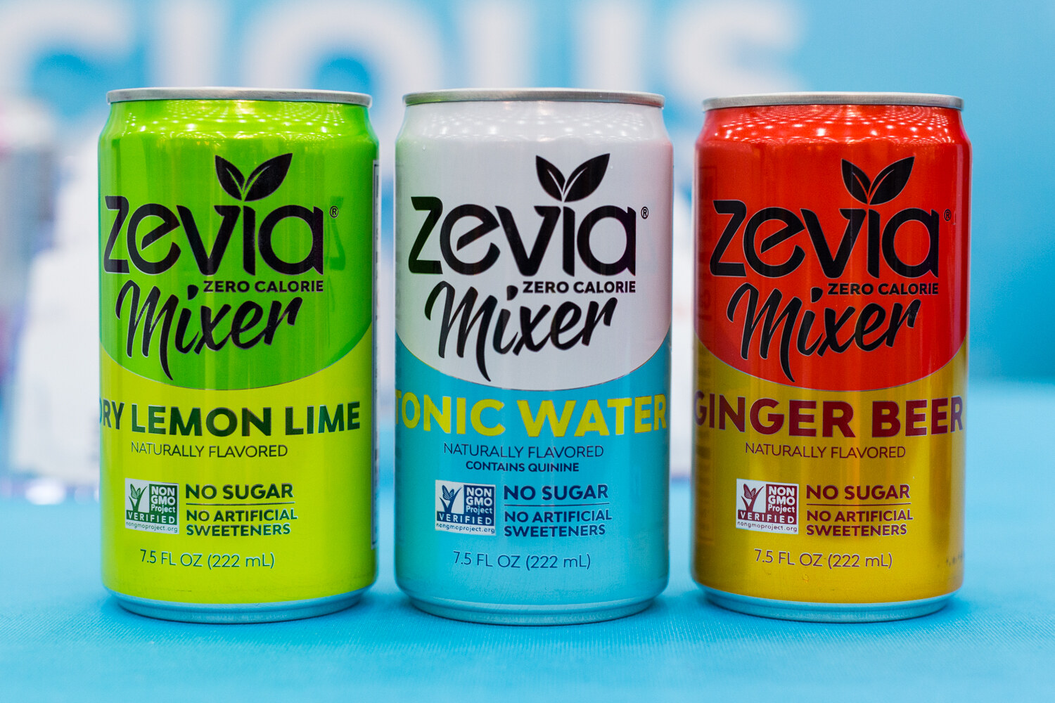Three Zevia Mixer cans in green, white and red on a blue tabletop.