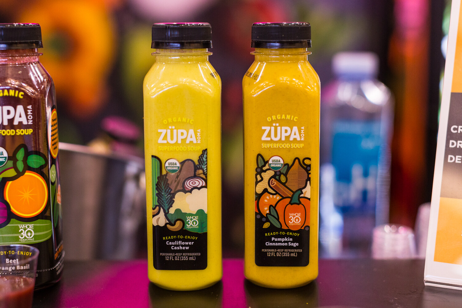 Two yellow bottles of Zupa Noma soup labeled Cauliflower Cashew and Pumpkin Cinnamon Sage beside a partly visible beet bottle.