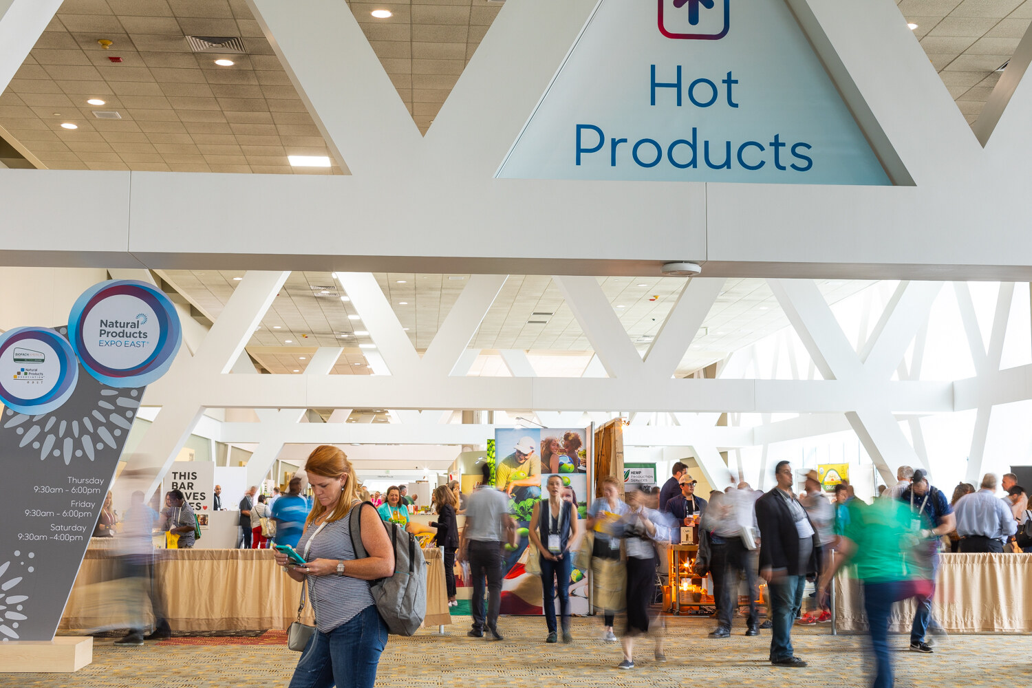 A crowd of badge-wearing attendees walking under a white lattice ceiling and a Hot Products sign, with show hours posted at left.