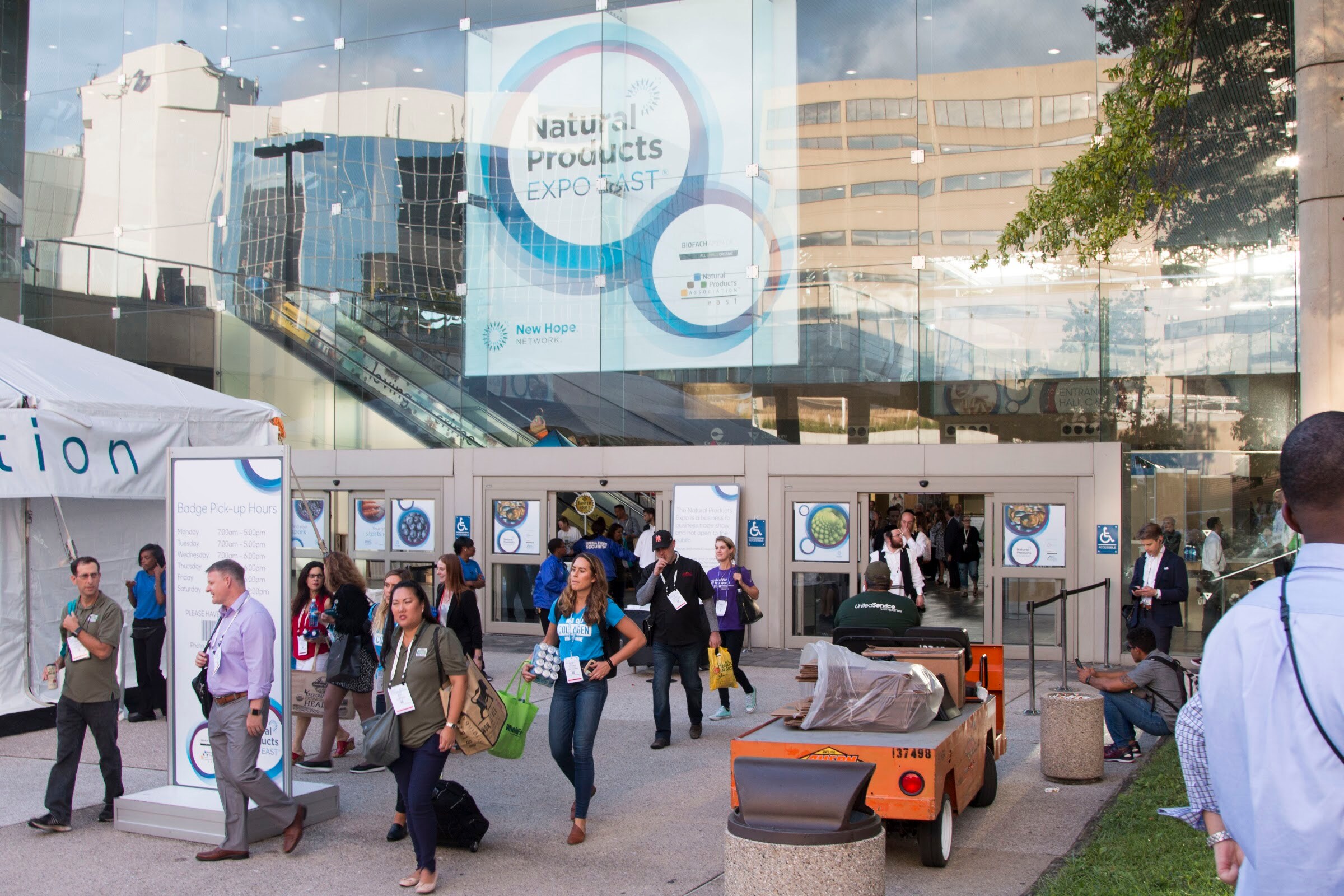 People walking in and out of glass doors below a large Natural Products Expo East banner, with a utility cart nearby.