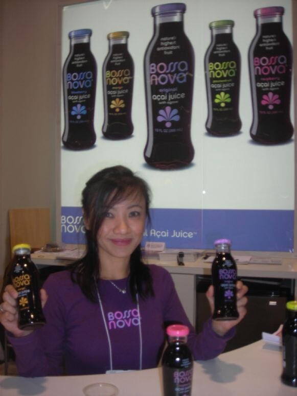 A smiling woman in a purple Bossa Nova shirt holds two bottles of açaí juice. A large backlit poster of Bossa Nova açaí juice bottles hangs behind her.