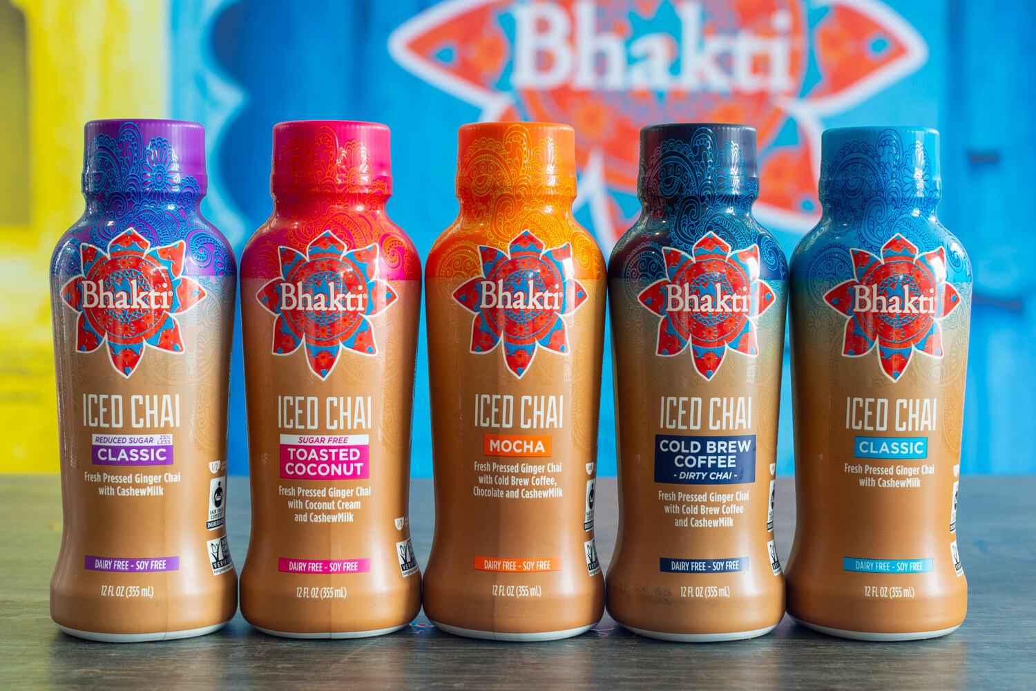 Five Bhakti iced chai bottles with mandala-style labels in front of a colorful booth backdrop.
