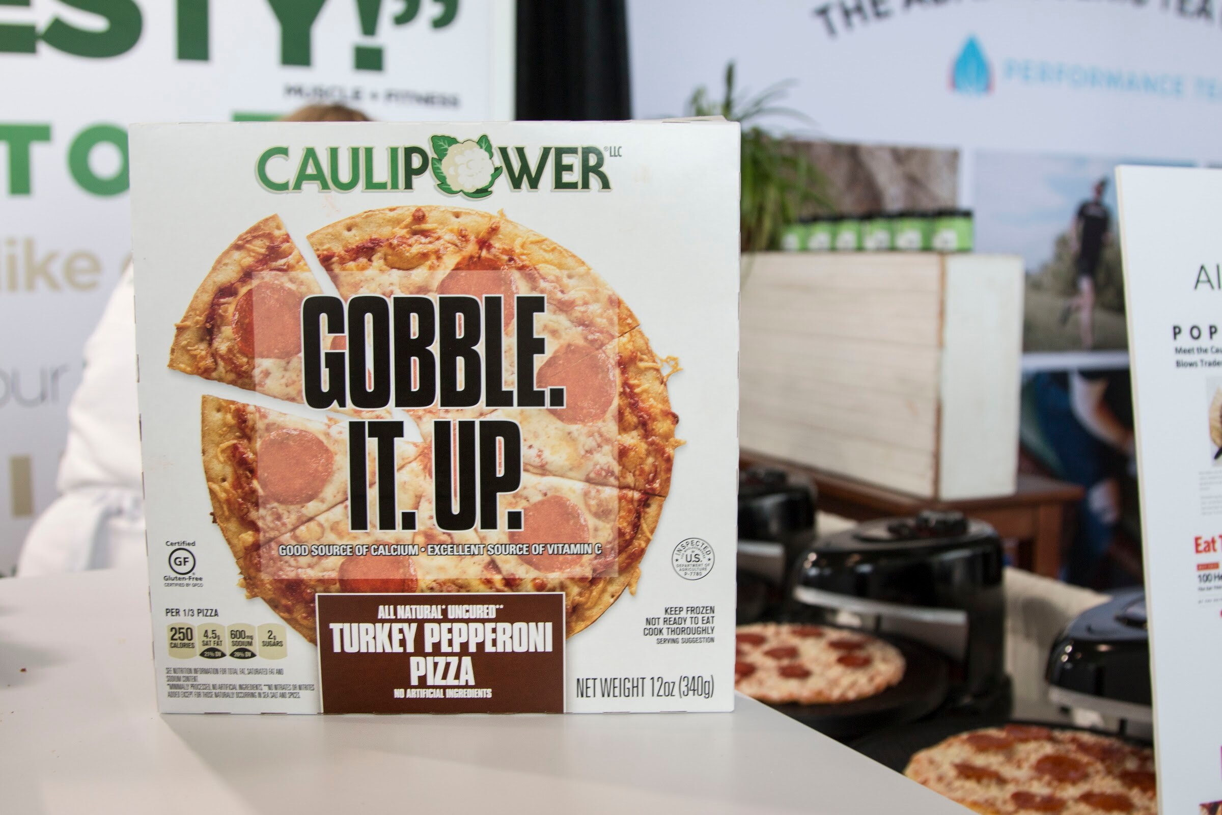 A large Caulipower pizza box on a white table with booth signage behind it.