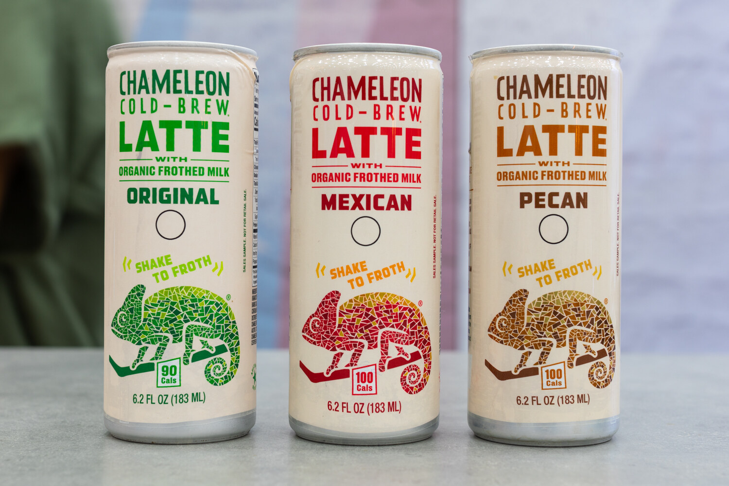 Three cream-colored Chameleon Cold-Brew Latte cans with mosaic chameleon art.