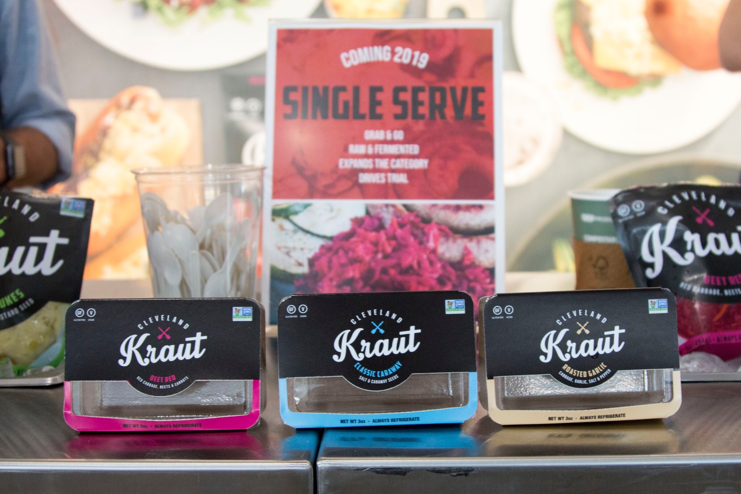 Three Cleveland Kraut cups in black, pink, blue and cream beside a red "Single serve" sign.