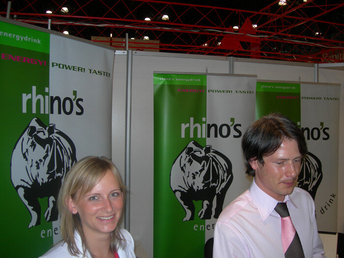 A smiling blonde woman and a dark-haired man in a pink shirt and tie stand in front of green and white rhino's energy drink banners showing a rhinoceros.
