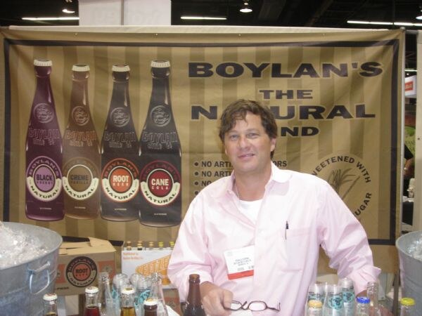 A man in a pink shirt stands behind a counter at the Boylan's booth, with a banner of Boylan's soda bottles behind him and iced buckets of bottles beside him.