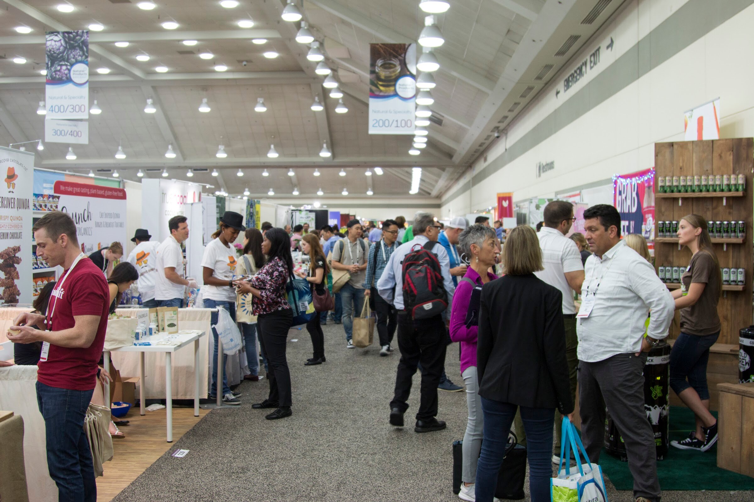 A packed carpeted aisle lined with booths and hanging banners, with people in conversation.