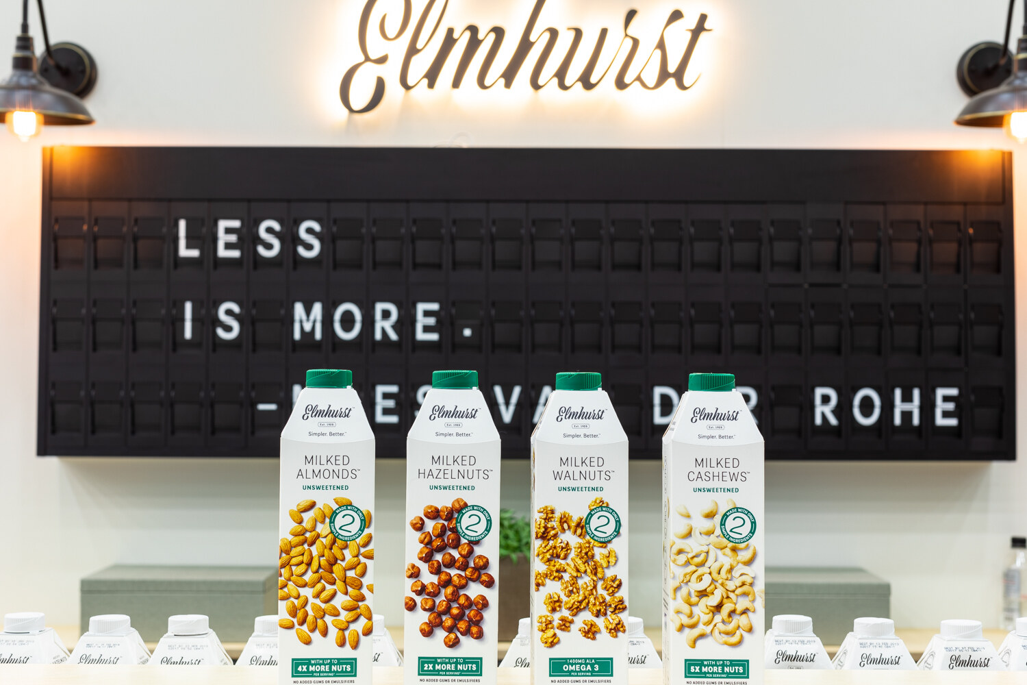 Four Elmhurst cartons in front of a black letterboard reading "Less is more" under an Elmhurst light-up sign.