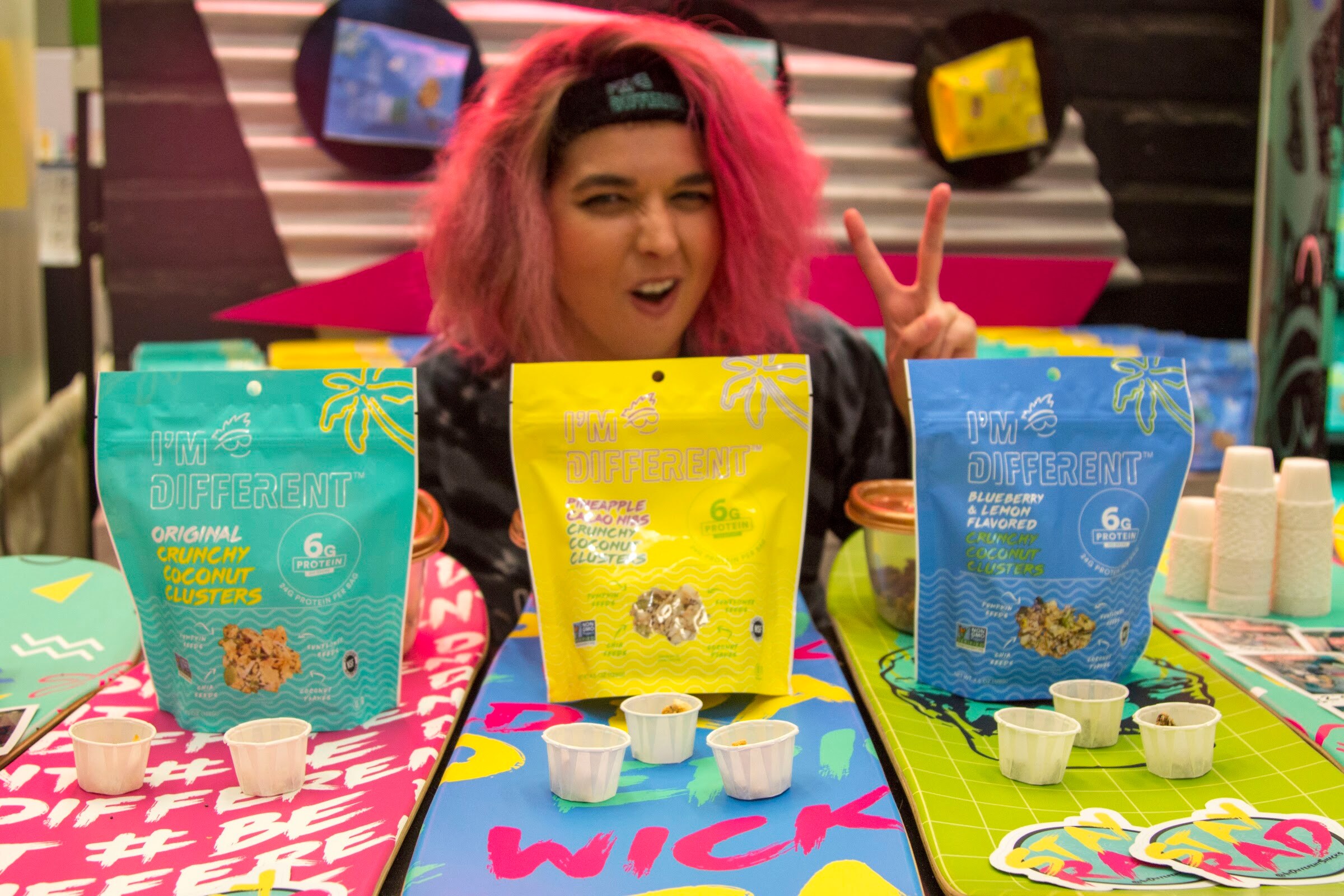 A smiling person with pink hair behind three bags of I'm Different clusters on a colorful table.
