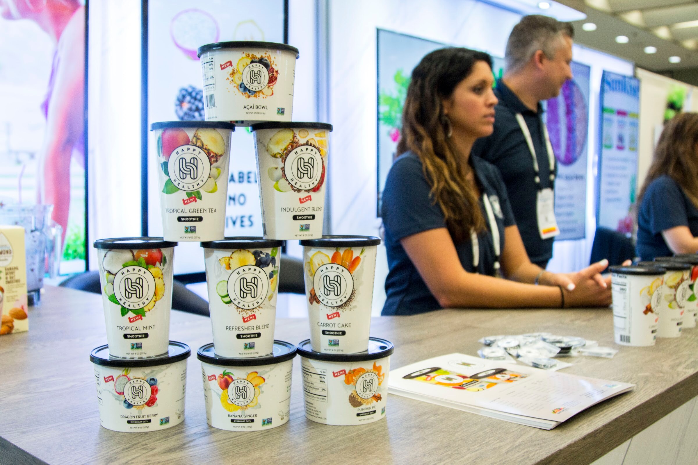 Two exhibitors with badges seated at a counter behind stacked white smoothie cups.