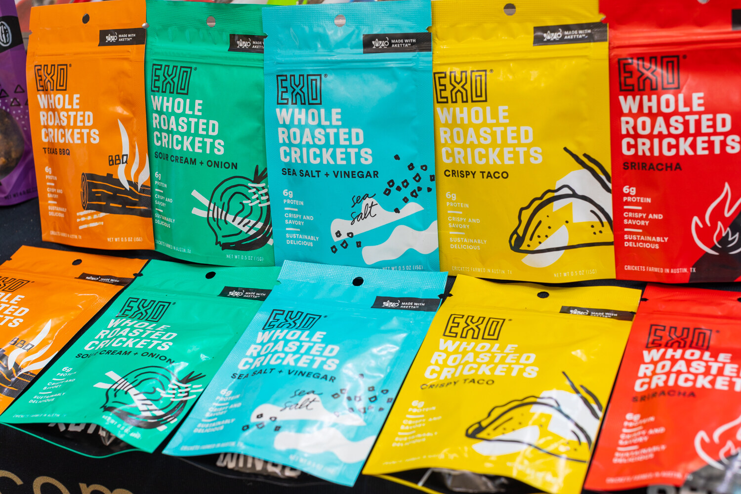 Rows of brightly colored Exo whole roasted cricket snack bags on a black table.