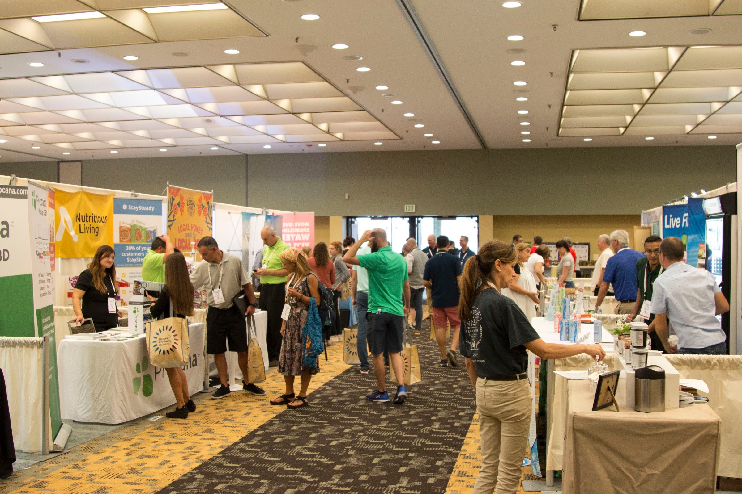 A carpeted exhibit hall aisle with booths on both sides and people sampling and talking.