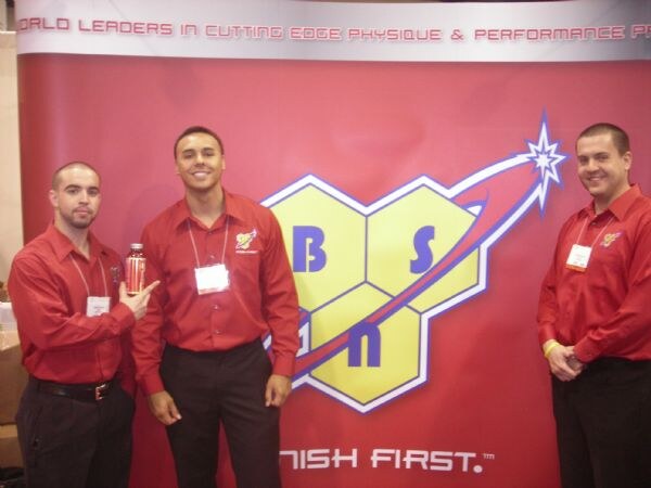 Three men in red shirts stand in front of a red BSN banner with a yellow hexagon logo. One man holds a bottle and another gestures toward it.
