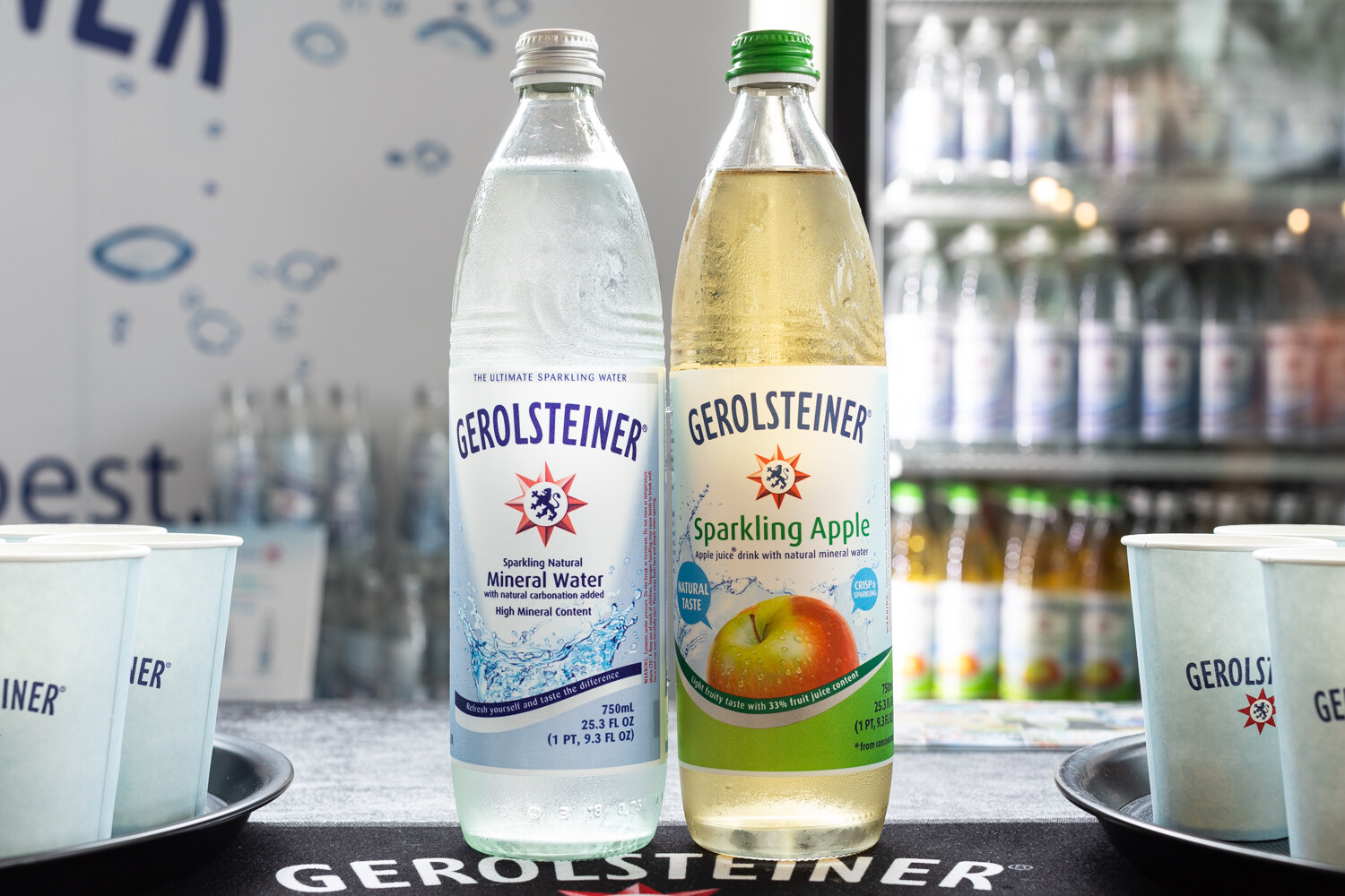 Two tall glass Gerolsteiner bottles, one clear and one pale green, on a black tray with paper cups.