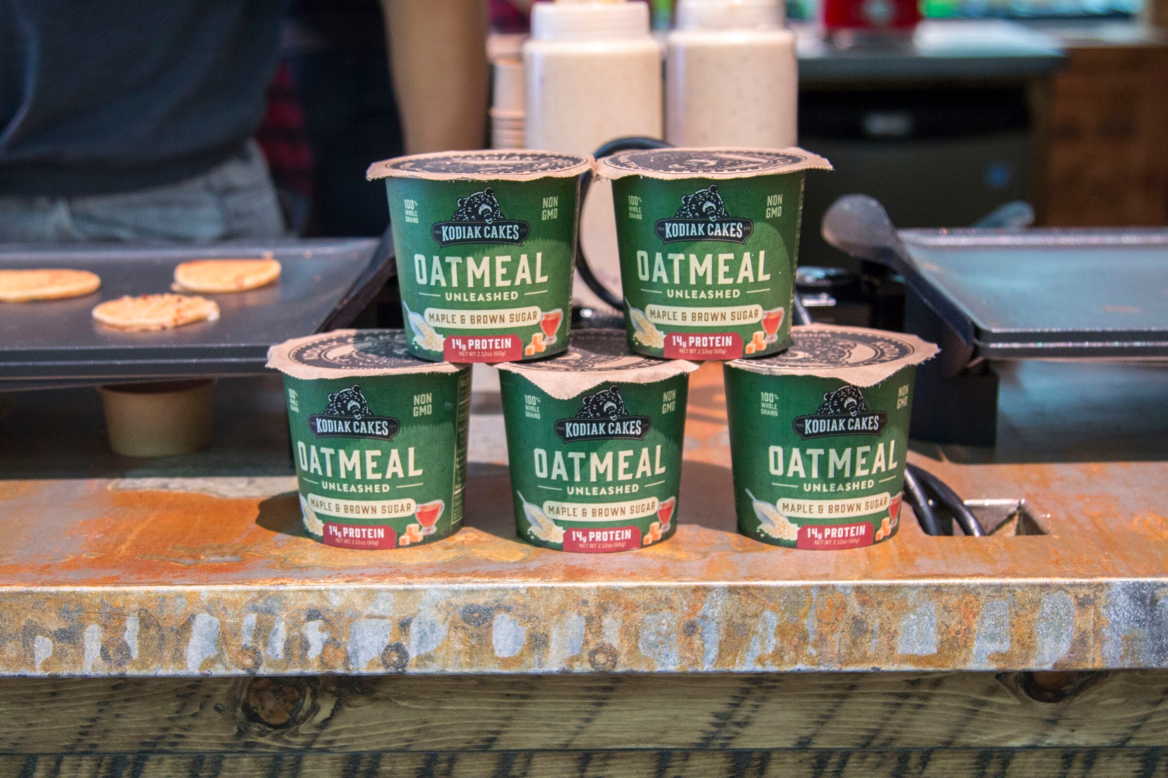 Five green Kodiak Cakes oatmeal cups stacked on a rustic counter next to a pancake griddle.