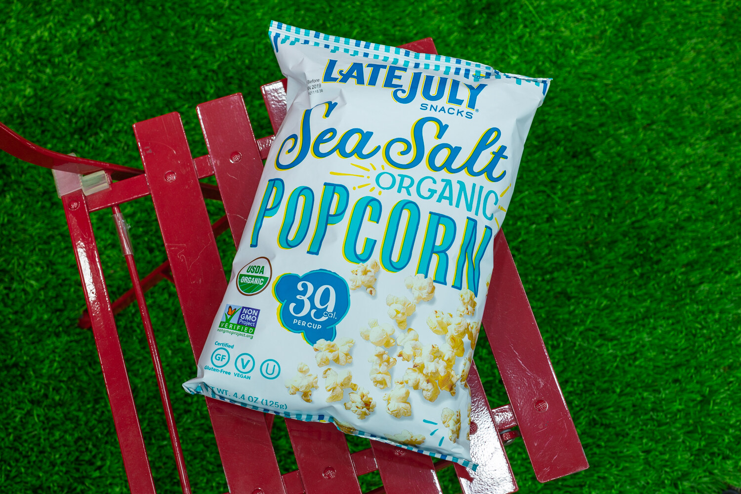 A Late July popcorn bag lying on a red bench against green turf.