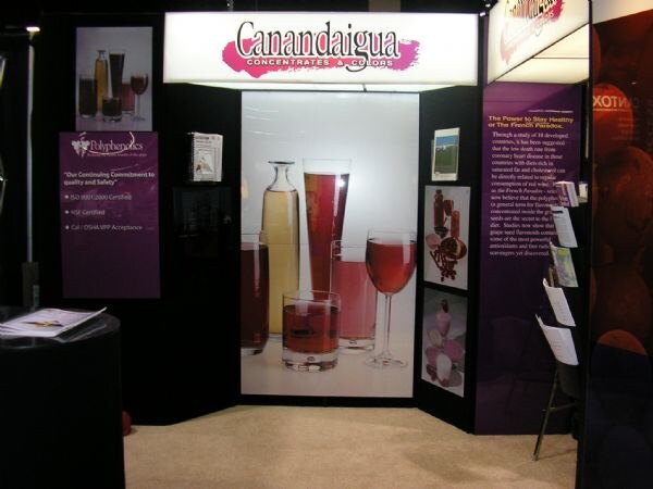 An empty trade-show booth with a lit Canandaigua Concentrates & Colors sign, a photo of glasses of wine and juice, and purple panels about polyphenols.