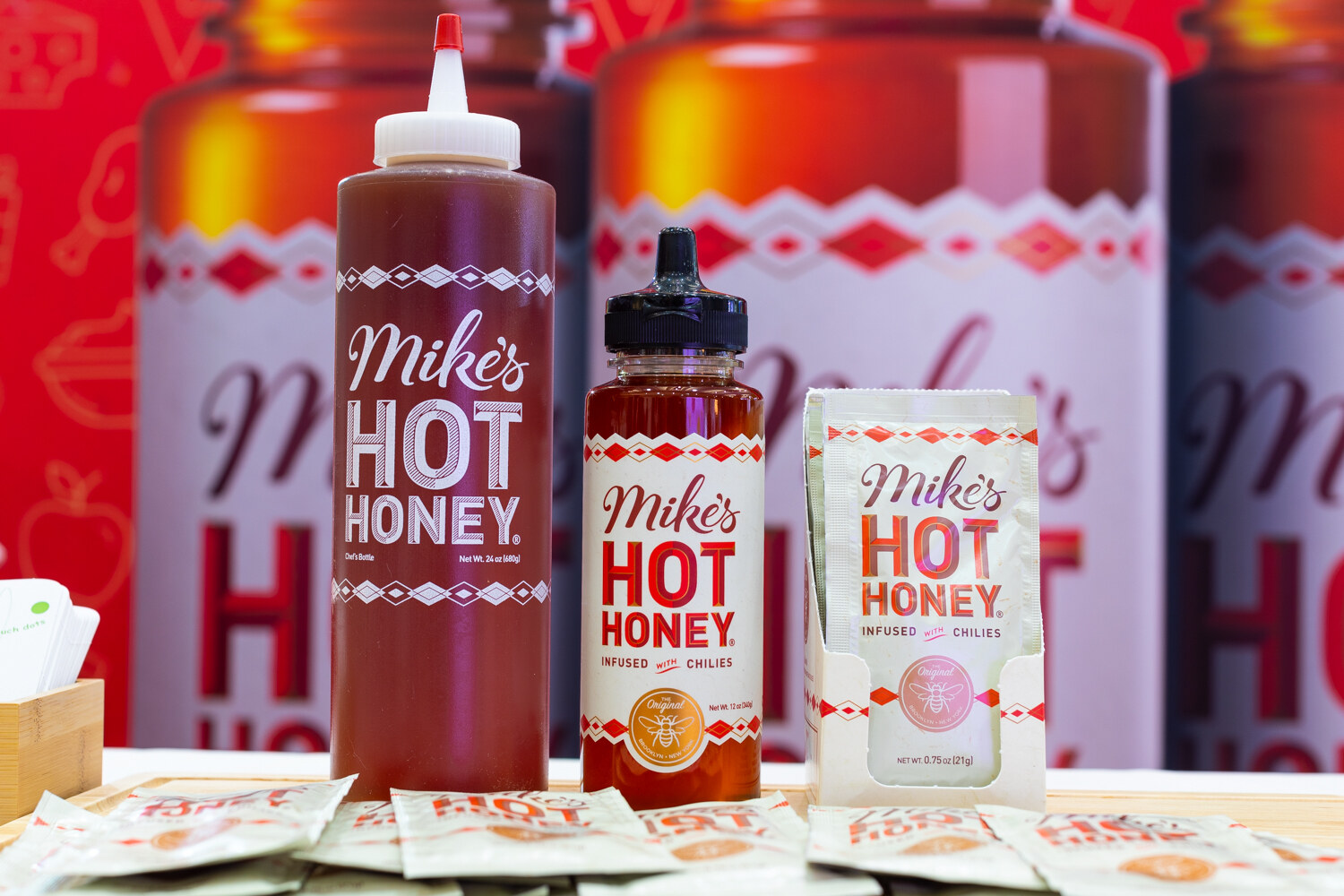 Three Mike's Hot Honey containers in front of a large red Mike's Hot Honey backdrop.