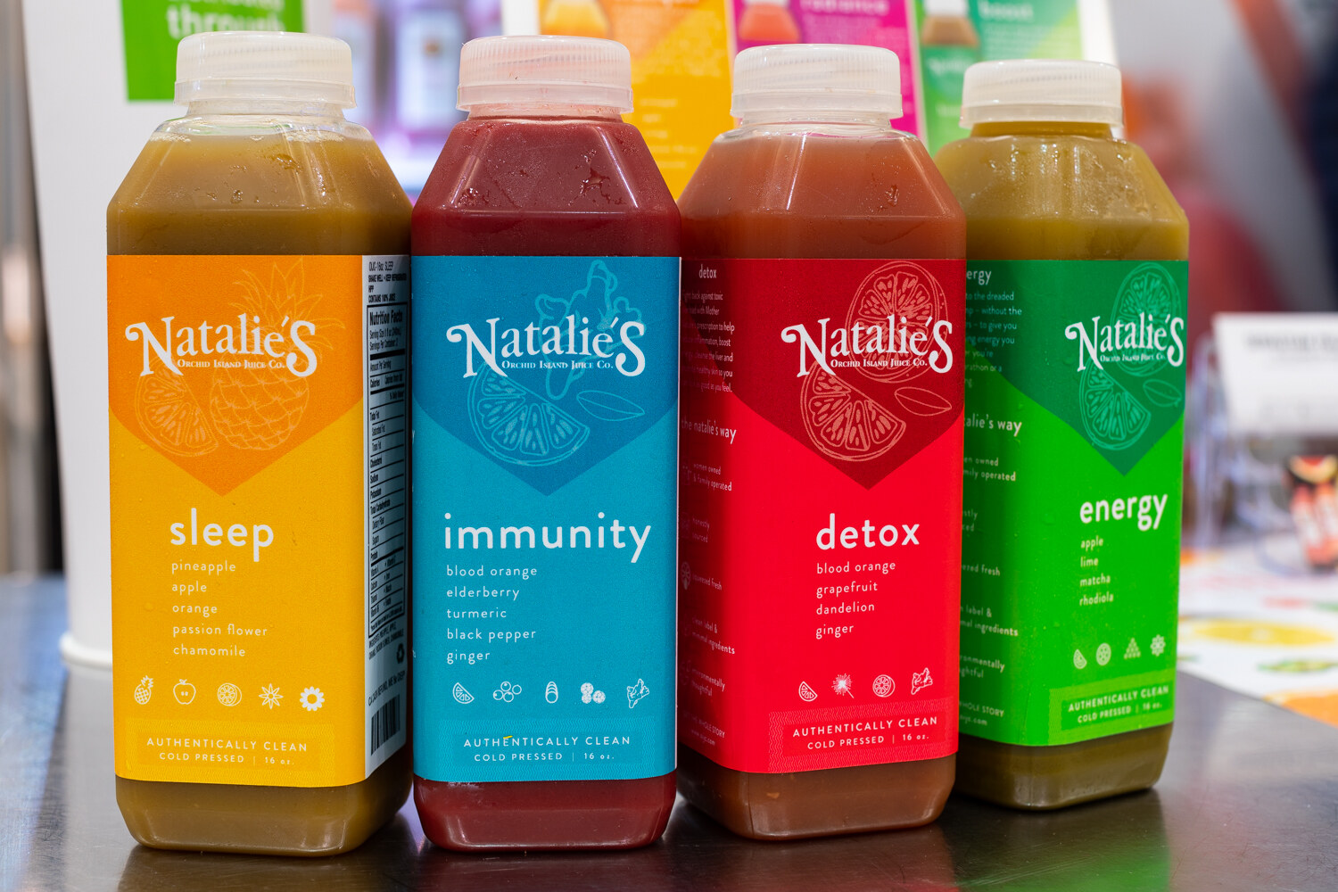 Four bottles of Natalie's juice in orange, blue, red and green labels lined up on a counter.