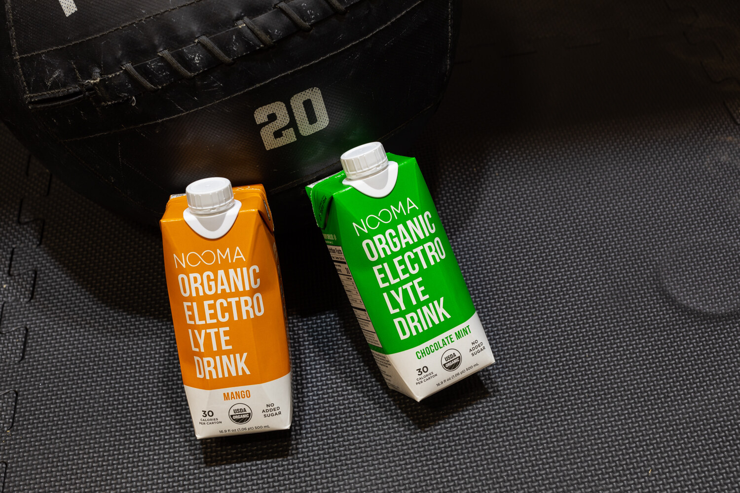 An orange and a green Nooma carton lying on a dark gym-mat surface beside a weight marked 20.