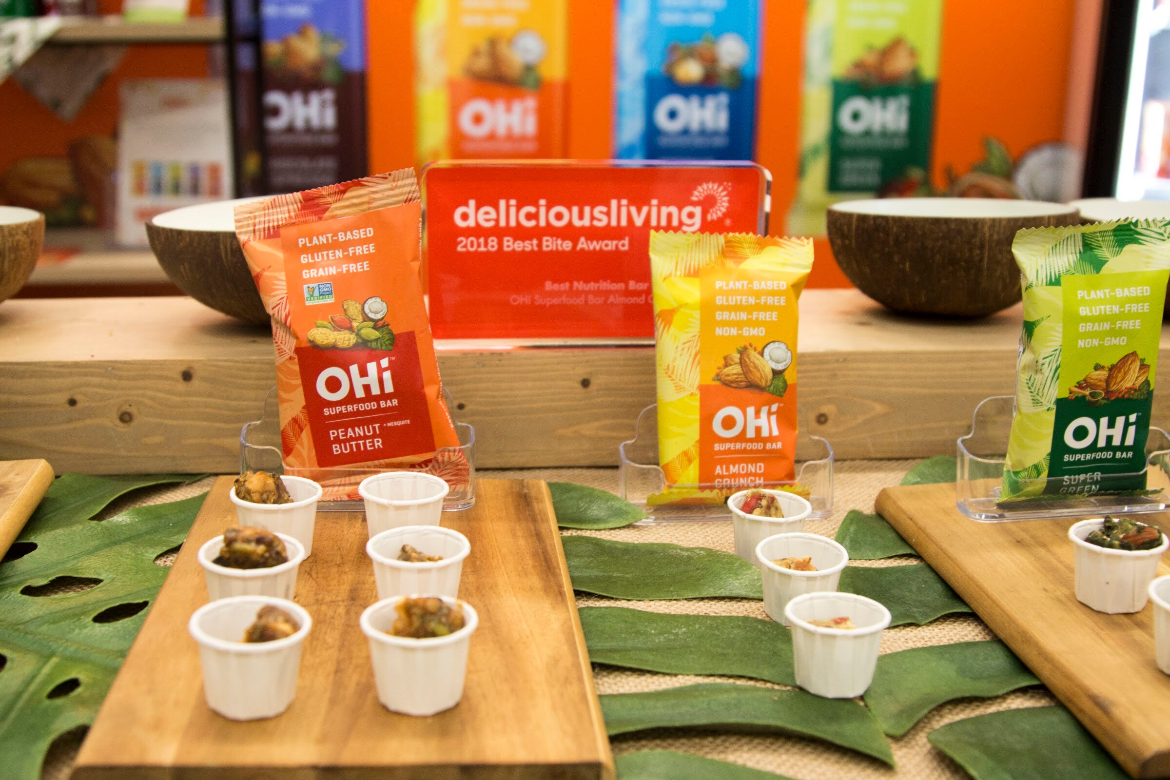 Three OHi bars on a wooden display with sample cups and a red Delicious Living award plaque.