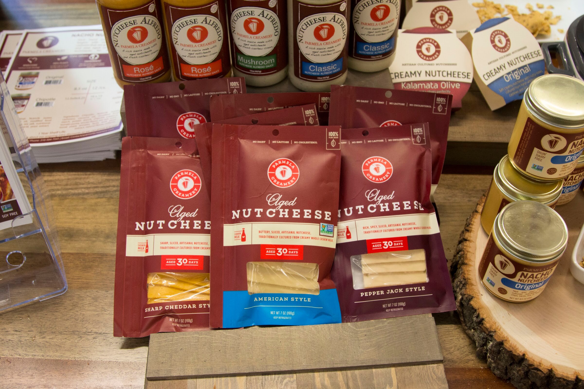 Burgundy Parmela Creamery nutcheese packages beside jars of nacho nutcheese on a wood slice.