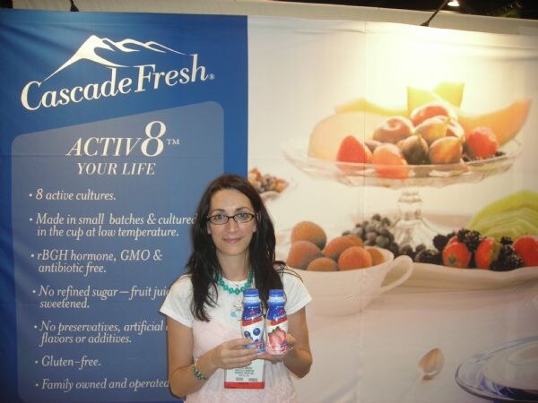 A woman with glasses and long dark hair holds two small bottles of drinkable yogurt in front of a blue and white Cascade Fresh Activ8 banner showing fruit.