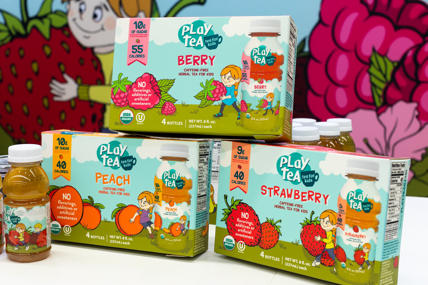 Three stacked Play Tea cartons with illustrated children and fruit in front of a strawberry mural.